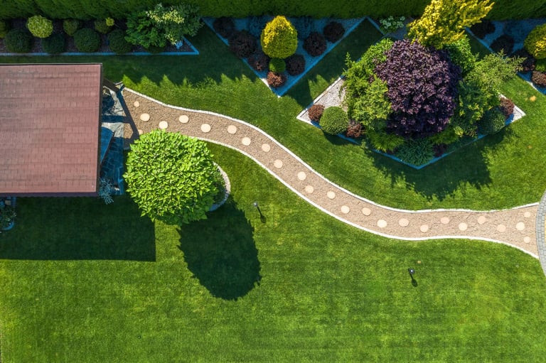 Aerial view of a vibrant garden featuring a winding stone path, ornamental shrubs, and well-manicured grass under bright sunlight, creating a serene atmosphere.