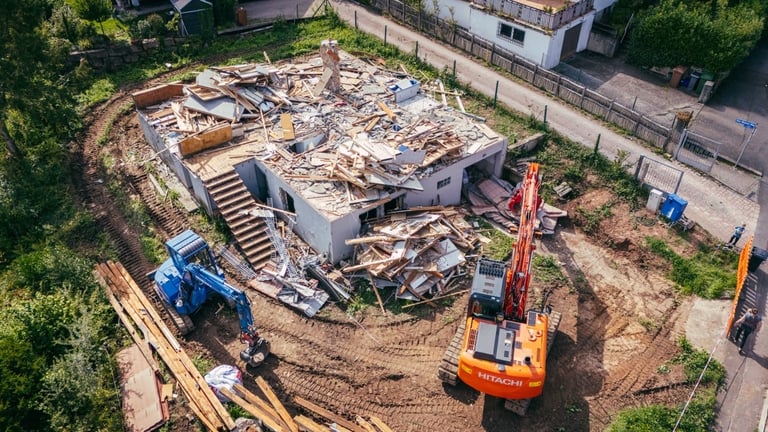 Aerial view of construction site with demolished building debris, heavy machinery, and orange excavator on disturbed soil surrounded by green vegetation.