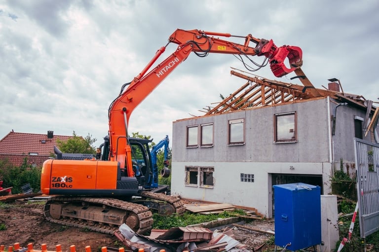 Orange excavator with extended arm demolishing a residential house with metal barrier in foreground