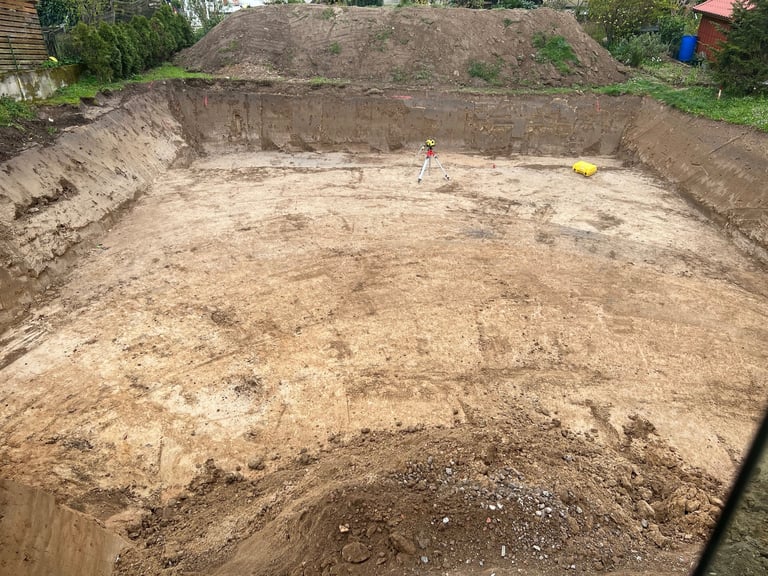 Large excavated pit with bare soil and earthen walls, surrounded by residential landscaping and a blue container visible in background