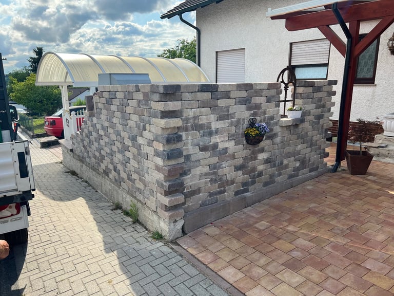 Gray patterned brick wall structure under construction in a residential driveway with paved pavers and house visible in background