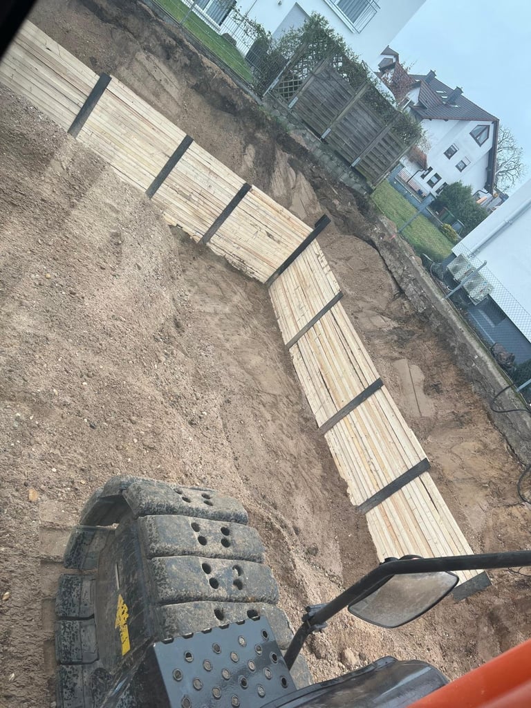 Construction site with newly laid wooden boards on sloped ground and shovel in foreground near residential buildings