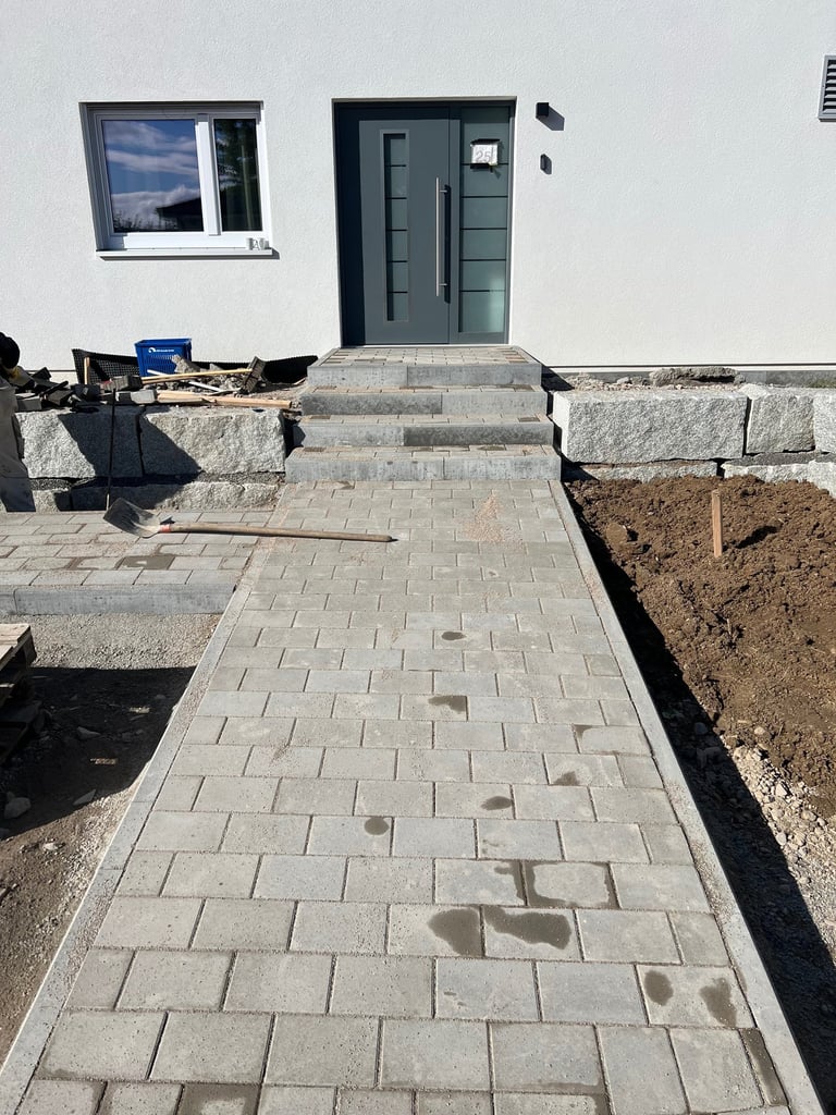 Modern house entrance under construction with paved walkway, dark double doors, window, and landscaping materials on sides