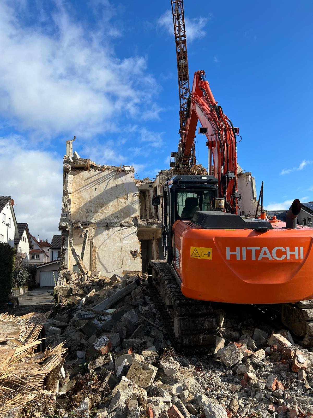 Roter Hitachi-Bagger reißt ein Betongebäude ab, mit Bauschutt im Vordergrund unter blauem Himmel