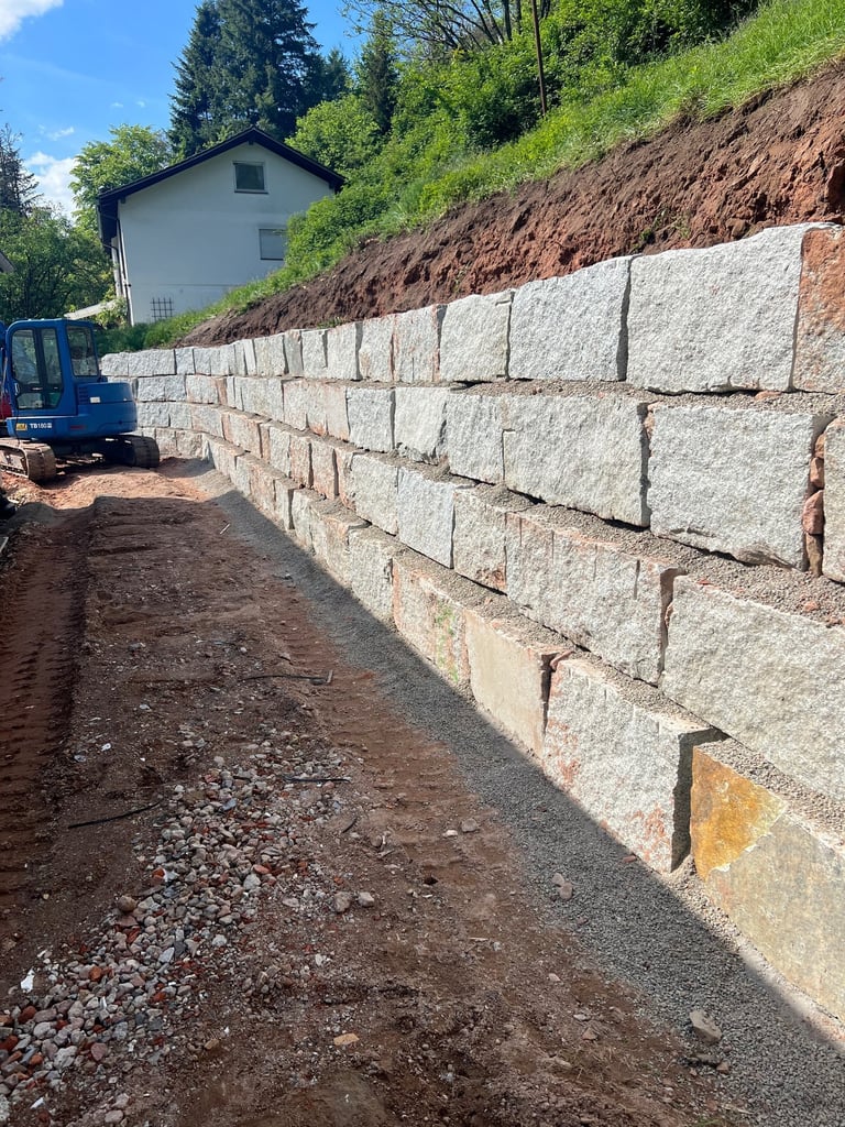 Large concrete retaining wall alongside dirt path with blue excavator and house in background surrounded by trees