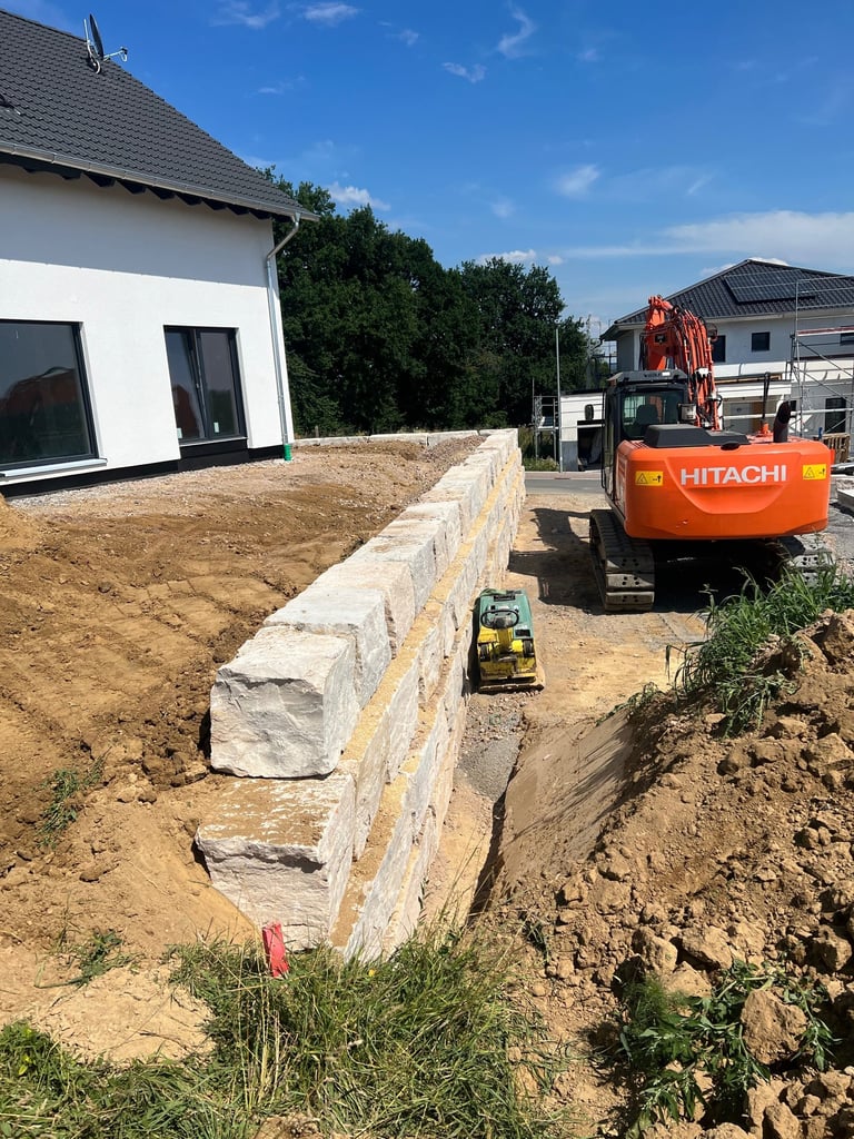 Construction site with concrete foundation blocks, orange Hitachi excavator, and white residential buildings under blue sky