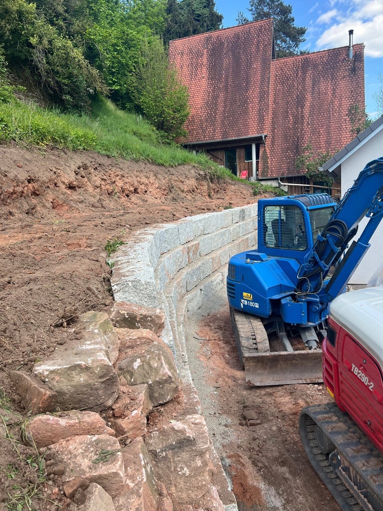 Blue excavator on a construction site with freshly dug earth, retaining wall, brick building, and vehicles parked nearby
