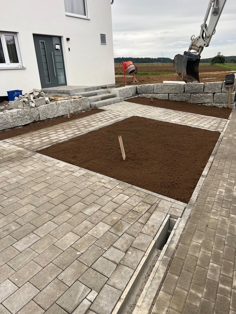 Construction site showing a foundation pit with brown soil surrounded by paver stones and concrete blocks next to a modern white house