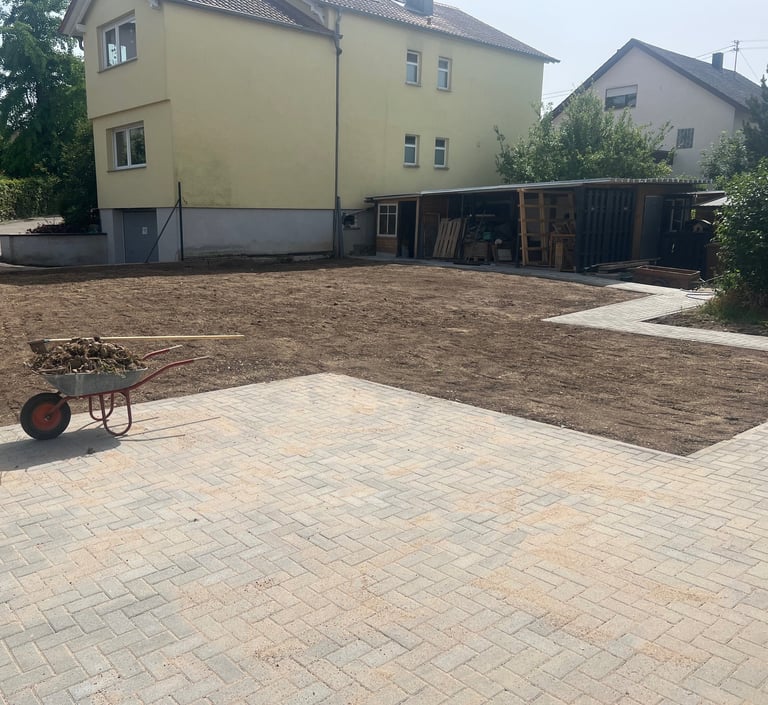Residential backyard with paved patio, mulched garden area, yellow house, storage shed, and wheelbarrow in foreground