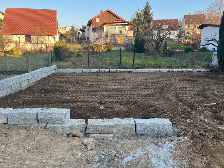 Prepared garden plot with concrete borders surrounded by fence and residential buildings in rural village setting